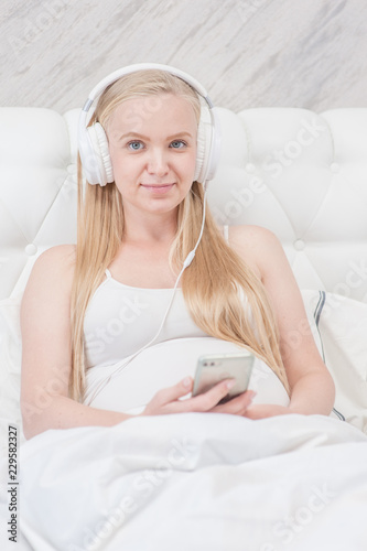 Happy pregnant woman listening to music on headphones on bed at home