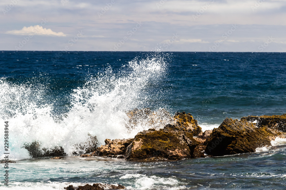 Naklejka premium Wave crashing on dark volcanic rock at Black Sand Beach (Punaluuu), on the Big Island of Hawaii. White spray rises in the air; Bue pacific ocean, sky and clouds are in the background. 