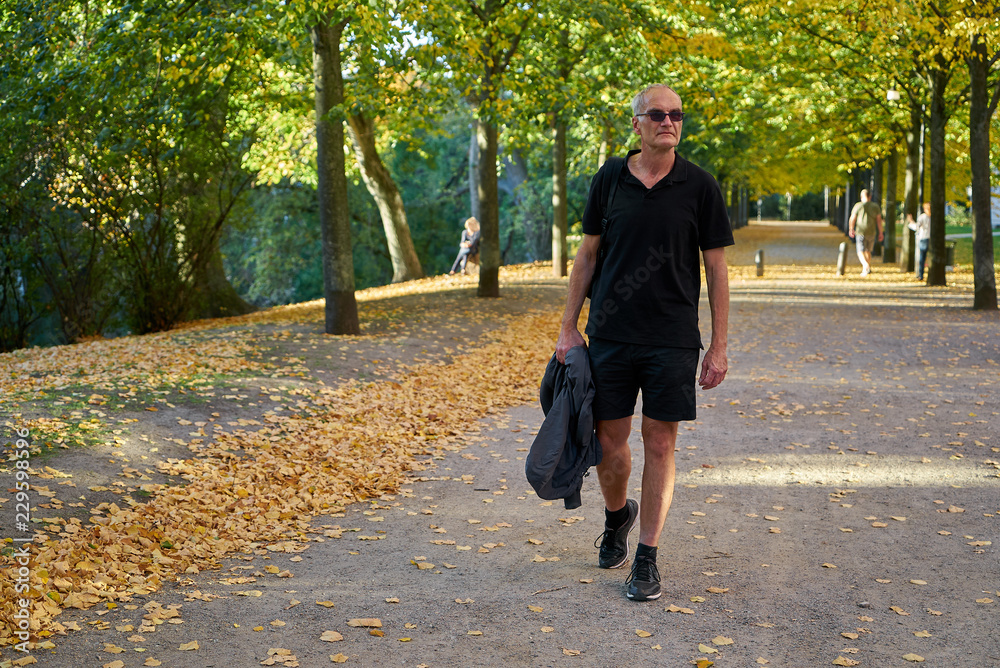 Fototapeta premium attraktiver europäischer Mann mittleren Alters mit kurzen Haaren, Sonnenbrille, schwarzem T-Shirt und Shorts spaziert durch einen herbstlichen Park
