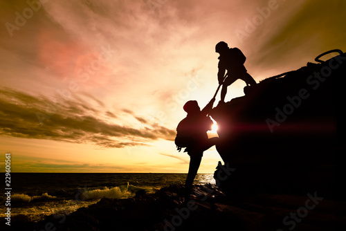 Silhouette of couple helping each other while climbing up on rocks beach in sunset. The concept of aid. photo