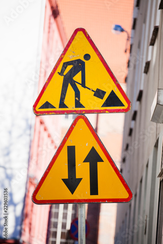 A yellow warning road sign indicating the repair work on a section of the road and under it a second road sign indicating movement in two directions