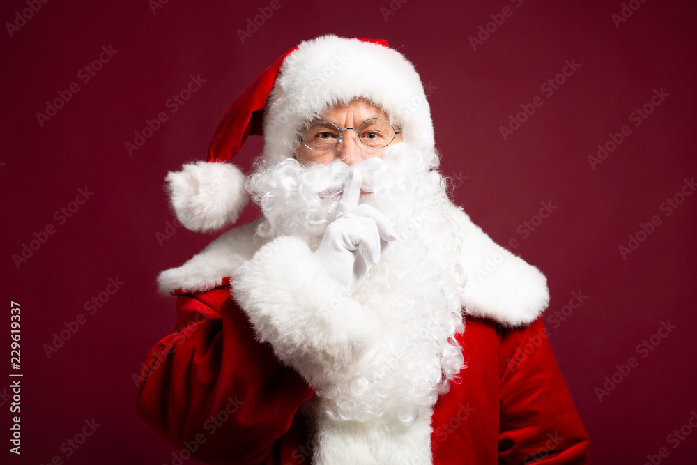 Portrait of man in Santa Clause costume looking at camera and showing silence sign on red background, Christmas and New year concept Portrait of man in Santa Clause costume looking at camera and showing silence sign on red background, Christmas and New year concept