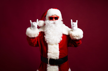 Stylish man in Santa Clause costume and sunglasses looking at camera and showing rock sign gesture on red background, Christmas and New year concept
