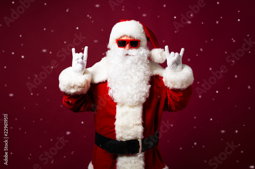 Stylish man in Santa Clause costume and sunglasses looking at camera and showing rock sign gesture on red background, Christmas and New year concept