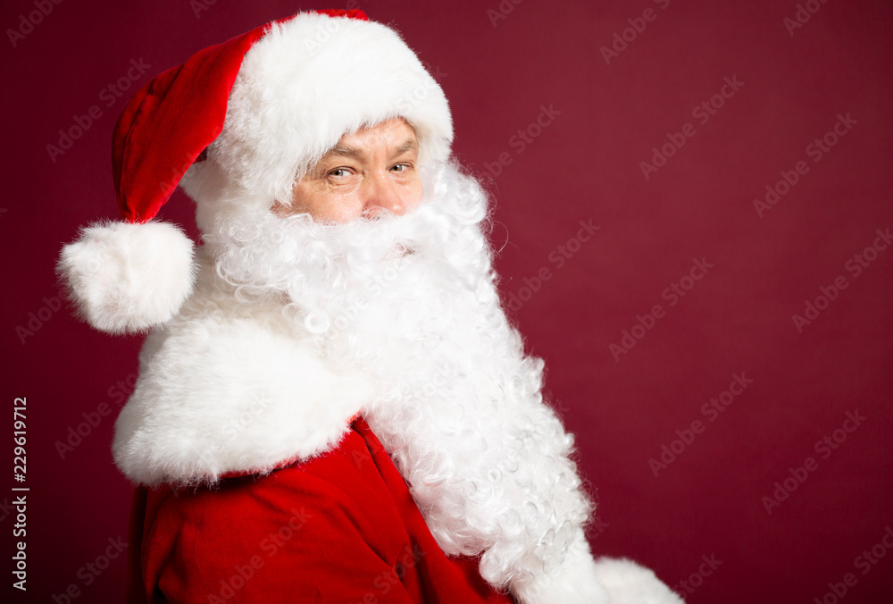 Side view of of man in Santa Clause costume looking at camera while posing on red background, Christmas and New year concept