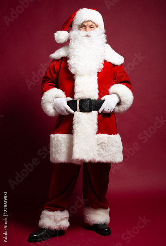 Man in Santa Clause costume holding hands on brown belt while posing on red background, Christmas and New year concept