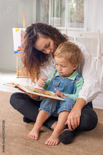 Young mother or nanny with small child boy sit on the floor on a rug in the room at home and read a book