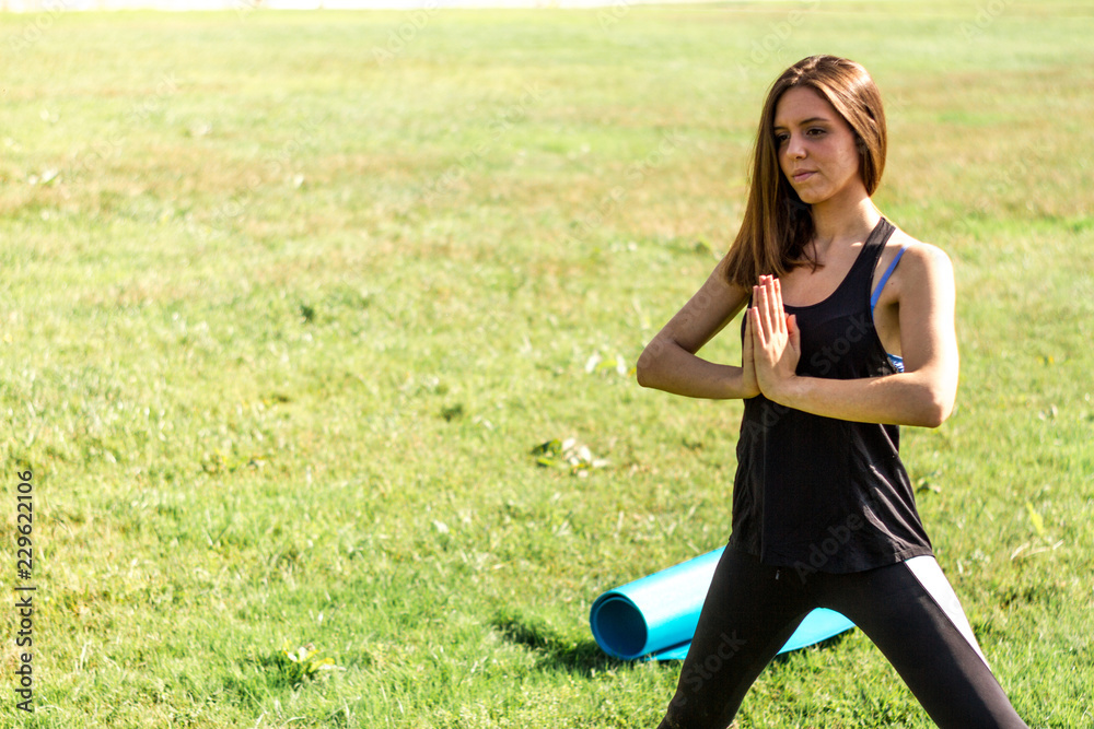 Young athletic woman practicing yoga in a green field with leafy grass next to a river. Young girl practicing postures and yoga exercises with an urban background Young athletic woman practicing yoga in a green field with leafy grass next to a river. Young girl practicing postures and yoga exercises with an urban background