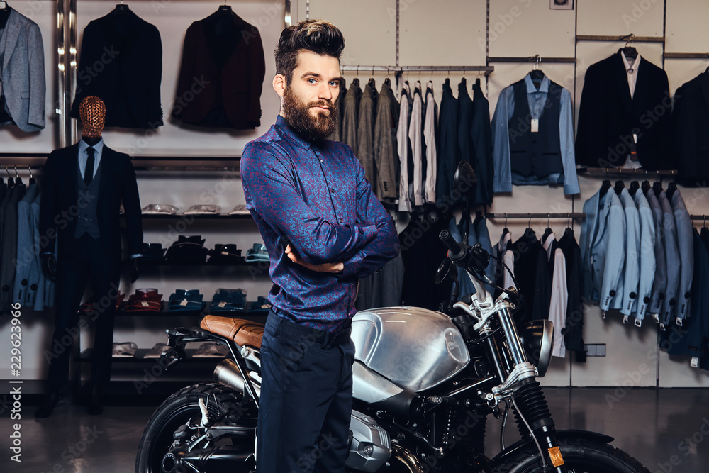 Fashionable man with full beard trendy dressed posing near retro sports motorbike at the men's clothing store.