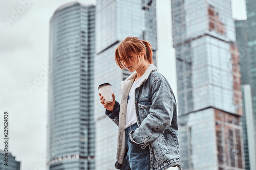 Stylish redhead hipster girl with tattoo on her face wearing denim jacket holding takeaway coffee in front of skyscrapers in Moskow city.