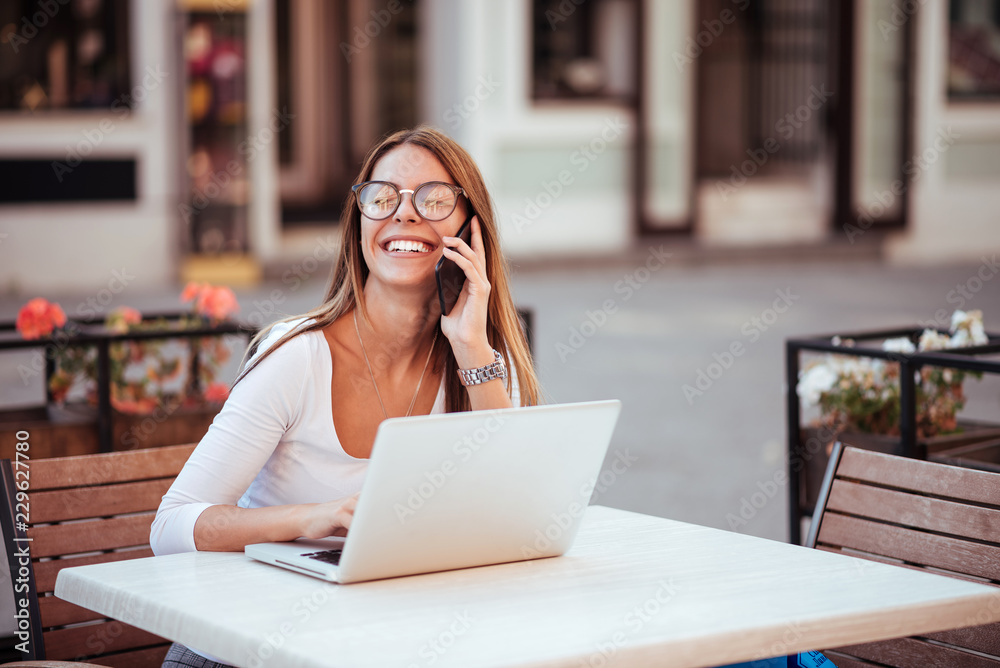 Girl with eyeglasses laughing while talking on the smatphone. Sitting with a laptop in the cafe outdoors. Girl with eyeglasses laughing while talking on the smatphone. Sitting with a laptop in the cafe outdoors.
