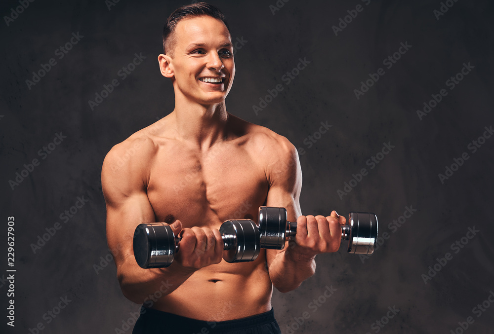 Young shirtless bodybuilder with muscular body doing exercise with dumbbells on dark background.