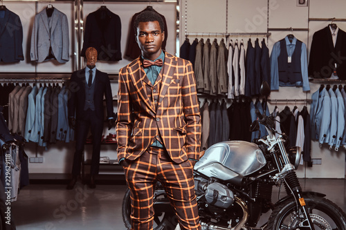 African-American man dressed in a trendy elegant suit posing with hands in pockets near retro sports motorbike at the men's clothing store. © Fxquadro