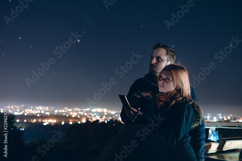 Loving couple is stand on the terrace by the wooden railing and make selfie on smartphone. look to distance, the lights of the night city. selective focus