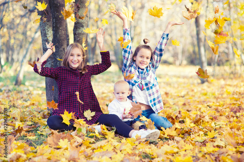Two cute smiling toddler girls and infant baby sitting in yellow foliage in park at warm sunny autumn day and drop leafs