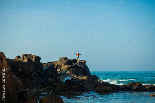 the girl stands on a rock and spread her hands to the sides and looks at the sea