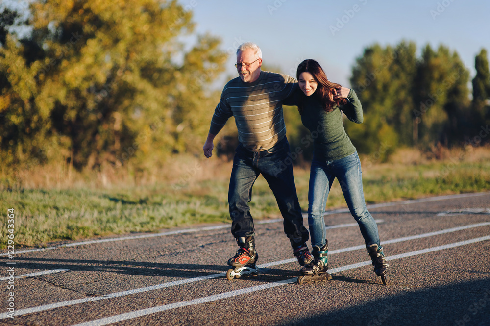 Parents and children spend time together. Older generation and sport. Active lifestyle of the pensioner. Happy old man riding on rollers with his daughter on the road in autumn park. Parents and children spend time together. Older generation and sport. Active lifestyle of the pensioner. Happy old man riding on rollers with his daughter on the road in autumn park.
