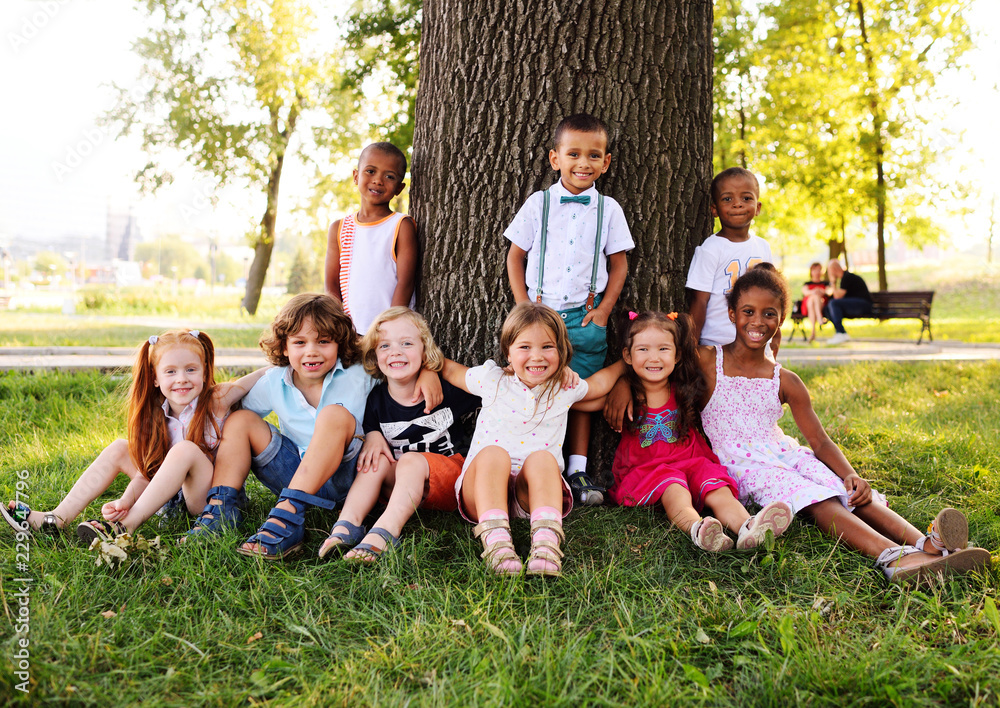 a group of small children in colorful clothes embracing sitting on the grass under a tree in a park laughing and smiling. June 1, Children's Day, vacation, friendship, friends, childhood. a group of small children in colorful clothes embracing sitting on the grass under a tree in a park laughing and smiling. June 1, Children's Day, vacation, friendship, friends, childhood.