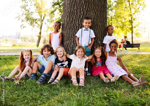 a group of small children in colorful clothes embracing sitting on the grass under a tree in a park laughing and smiling. June 1, Children's Day, vacation, friendship, friends, childhood.