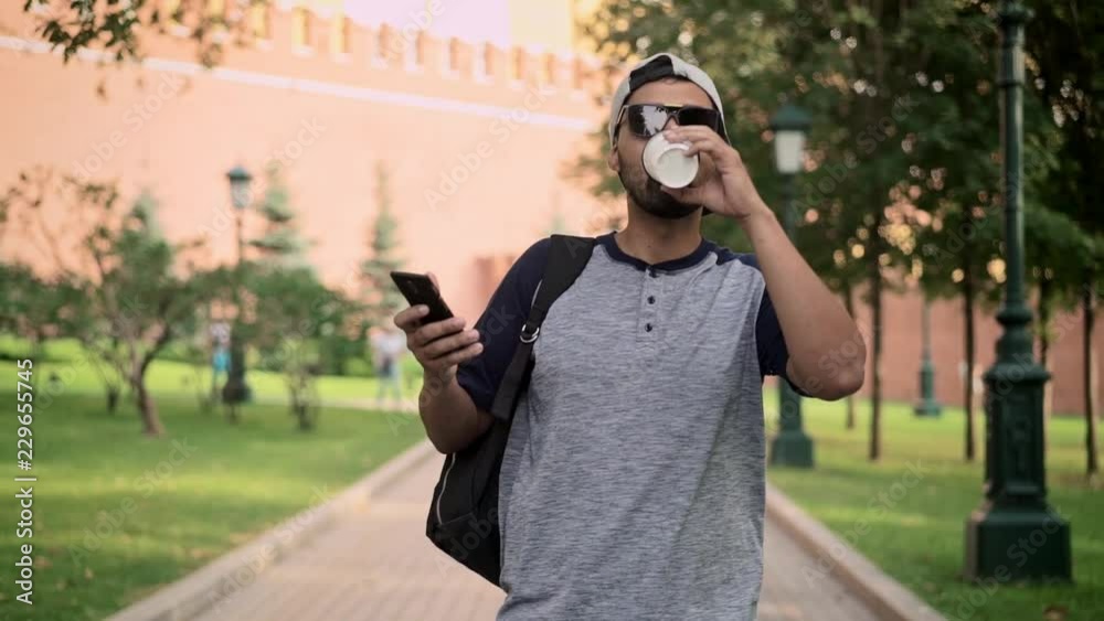 Cheerful young man wearing sunglasses and baseball cap walking near Kremlin, drinking his coffee and holding a smartphone. Tracking slow motion medium shot Cheerful young man wearing sunglasses and baseball cap walking near Kremlin, drinking his coffee and holding a smartphone. Tracking slow motion medium shot