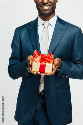 Vertical detail of man in suit and tie smiling and holding expensive golden gift with red ribbon, isolated on white studio background