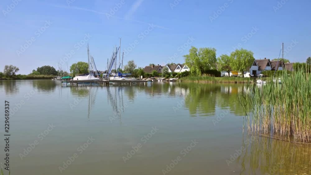 Morning time-lapse of Lake Neusiedlersee in Austria. Picturesque holiday homes in terraced settlement of Seepark Weiden under sunny blue sky. Reed grows on the shore. Boats are at boat dock. Morning time-lapse of Lake Neusiedlersee in Austria. Picturesque holiday homes in terraced settlement of Seepark Weiden under sunny blue sky. Reed grows on the shore. Boats are at boat dock.