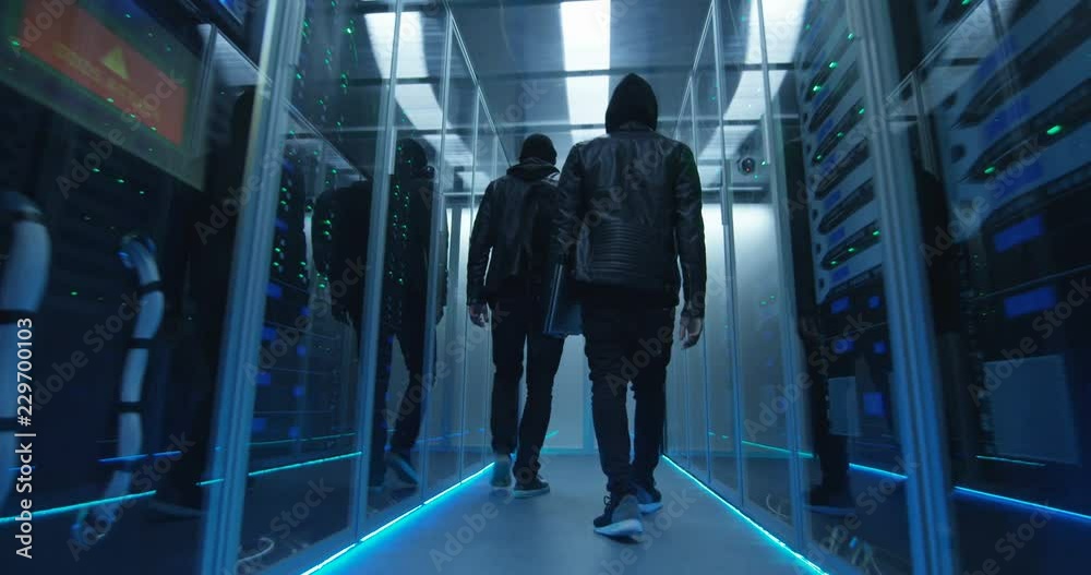 Wide shot from behind of a two hackers walking through corporate data center with rows of working rack servers