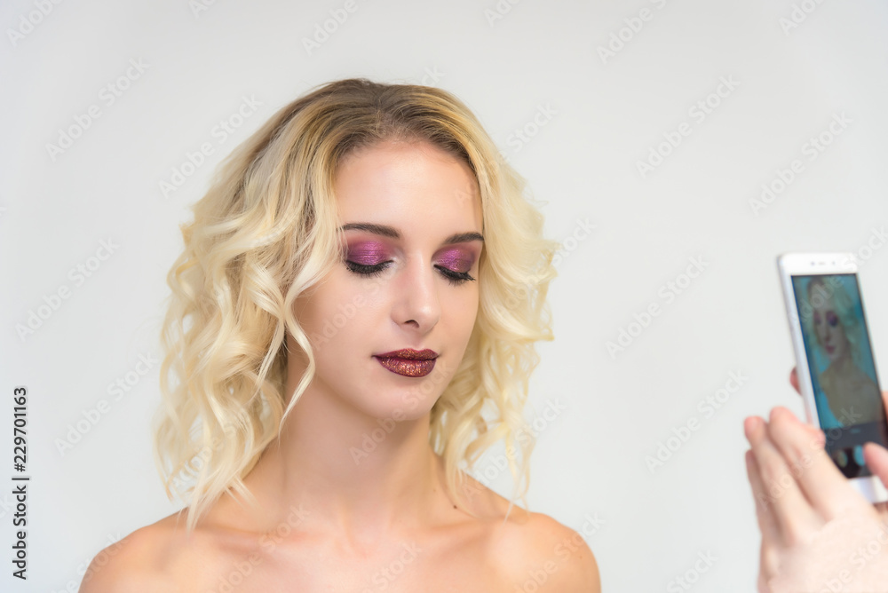 Beauty portrait of a beautiful blonde girl on a white background with a perfect make-up makes selfie on a smartphone Beauty portrait of a beautiful blonde girl on a white background with a perfect make-up makes selfie on a smartphone