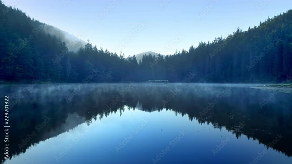 Clear blue mountain lake covered with low fog Aerial flight from water surface level Reflections of sky and mountains falling in water