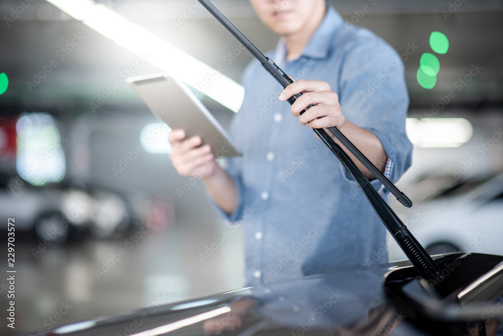 Young Asian auto mechanic holding digital tablet checking windshield wiper in auto service garage. Mechanical maintenance engineer working in automotive industry. Automobile servicing and repair