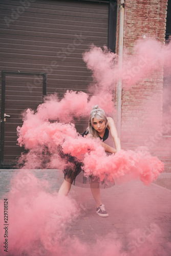 attractive young woman dancing in pink smoke and looking at camera on street