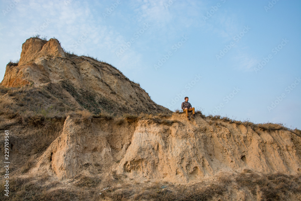 Young male hipster traveler relaxing on the cliff near sea against sunset
