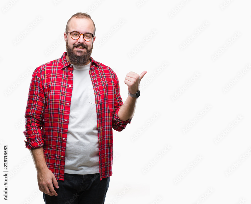Young caucasian hipster man wearing glasses over isolated background smiling with happy face looking and pointing to the side with thumb up. Young caucasian hipster man wearing glasses over isolated background smiling with happy face looking and pointing to the side with thumb up.