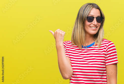 Young beautiful woman wearing sunglasses over isolated background smiling with happy face looking and pointing to the side with thumb up.