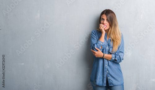 Young adult woman over grunge grey wall wearing denim outfit looking stressed and nervous with hands on mouth biting nails. Anxiety problem.
