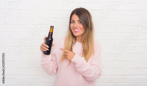 Yound adult woman over white brick wall holding beer very happy pointing with hand and finger