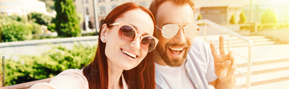 panoramic view of happy boyfriend and girlfriend in sunglasses looking at camera in city, man showing peace sign panoramic view of happy boyfriend and girlfriend in sunglasses looking at camera in city, man showing peace sign