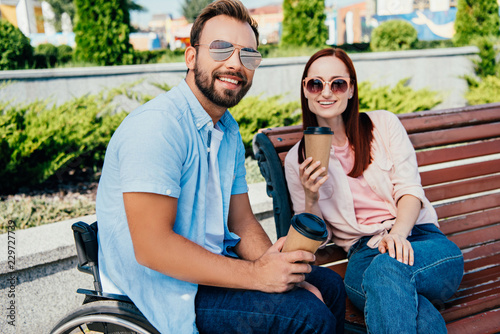 smiling handsome boyfriend in wheelchair and girlfriend looking at camera and holding disposable coffee cups on street