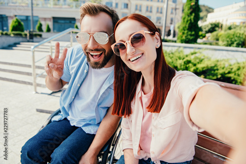camera point of view of happy boyfriend in wheelchair and attractive girlfriend looking at camera in city, man showing peace sign
