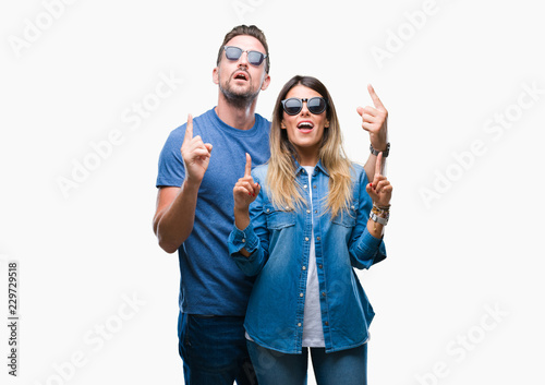 Young couple in love wearing sunglasses over isolated background amazed and surprised looking up and pointing with fingers and raised arms.