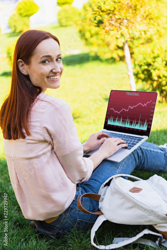 smiling attractive woman using laptop with traiding appliance in park and looking at camera photo