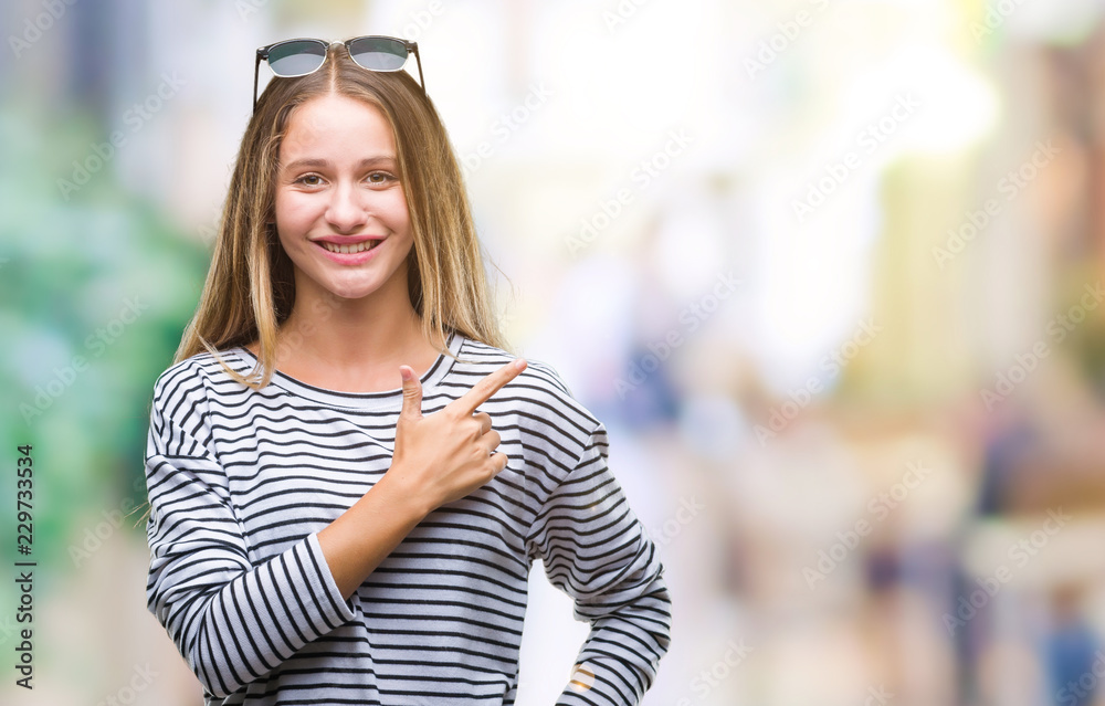 Young beautiful blonde woman wearing sunglasses over isolated background cheerful with a smile of face pointing with hand and finger up to the side with happy and natural expression on face