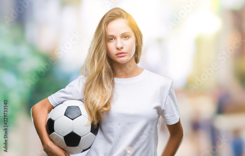 Young beautiful blonde woman holding soccer ball over isolated background with a confident expression on smart face thinking serious © Krakenimages.com