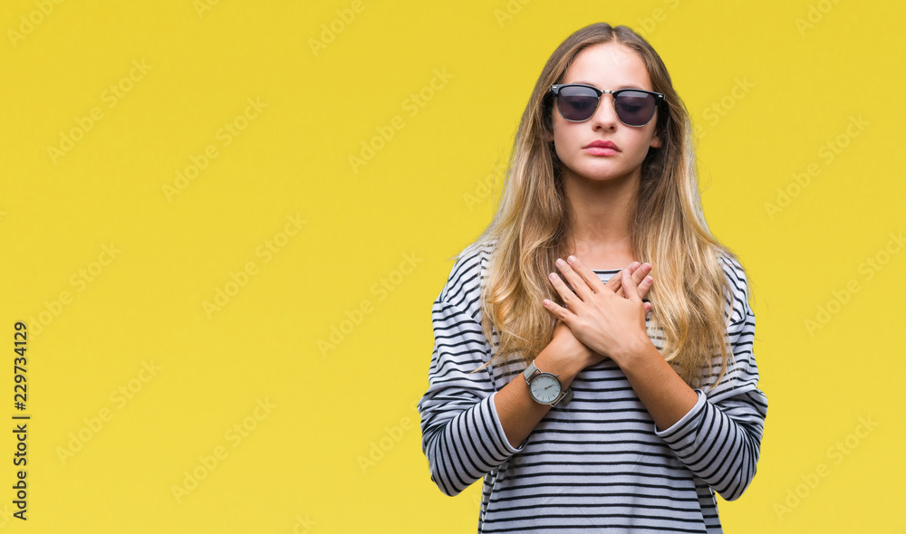Fototapeta premium Young beautiful blonde woman wearing sunglasses over isolated background smiling with hands on chest with closed eyes and grateful gesture on face. Health concept.