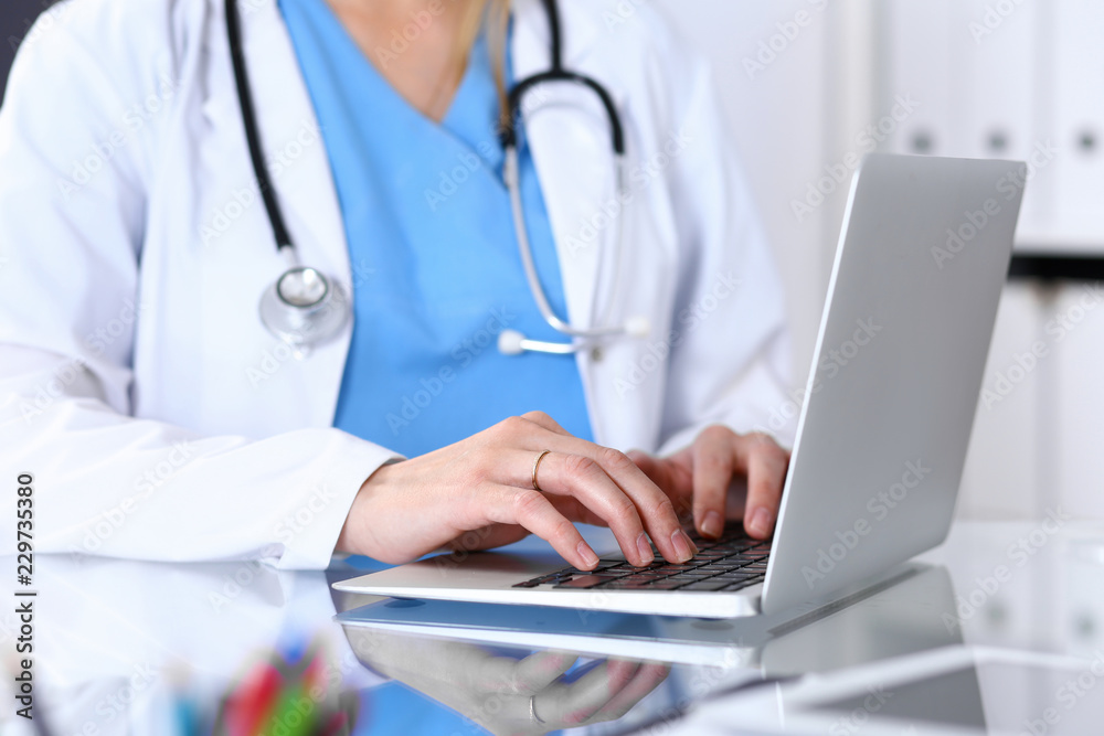 Doctor typing on laptop computer while sitting at the glass desk in hospital office. Physician at work. Medicine and healthcare concept Doctor typing on laptop computer while sitting at the glass desk in hospital office. Physician at work. Medicine and healthcare concept