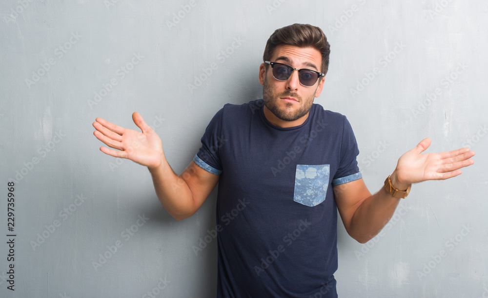 Fototapeta premium Handsome young man over grey grunge wall wearing sunglasses clueless and confused expression with arms and hands raised. Doubt concept.