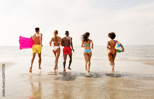 friendship, summer holidays and swimming concept - happy friends with floating mattress and inflatable ball running on beach