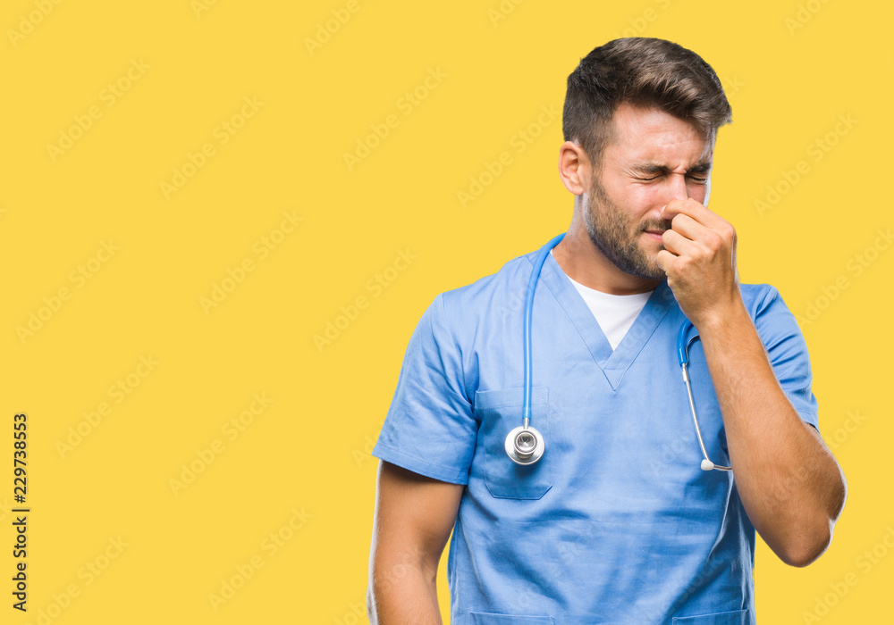 Young handsome doctor surgeon man over isolated background smelling something stinky and disgusting, intolerable smell, holding breath with fingers on nose. Bad smells concept. Young handsome doctor surgeon man over isolated background smelling something stinky and disgusting, intolerable smell, holding breath with fingers on nose. Bad smells concept.