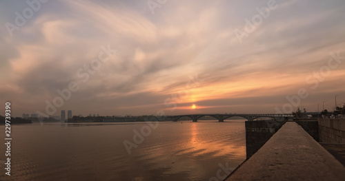 Wide angle landscape panorama of Dnipro River, embankment and Merto Bridge. Colorful vibrant sky reflected in the water. Foggy autumn morning during sunrise. Kyiv, Ukraine
