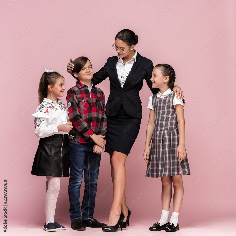 Cute smiling happy stylish children and female teacher on pink background. Beautiful stylish teen girls and boy standing together and posing at studio. Classic style. Kids fashion and emotions concept Cute smiling happy stylish children and female teacher on pink background. Beautiful stylish teen girls and boy standing together and posing at studio. Classic style. Kids fashion and emotions concept